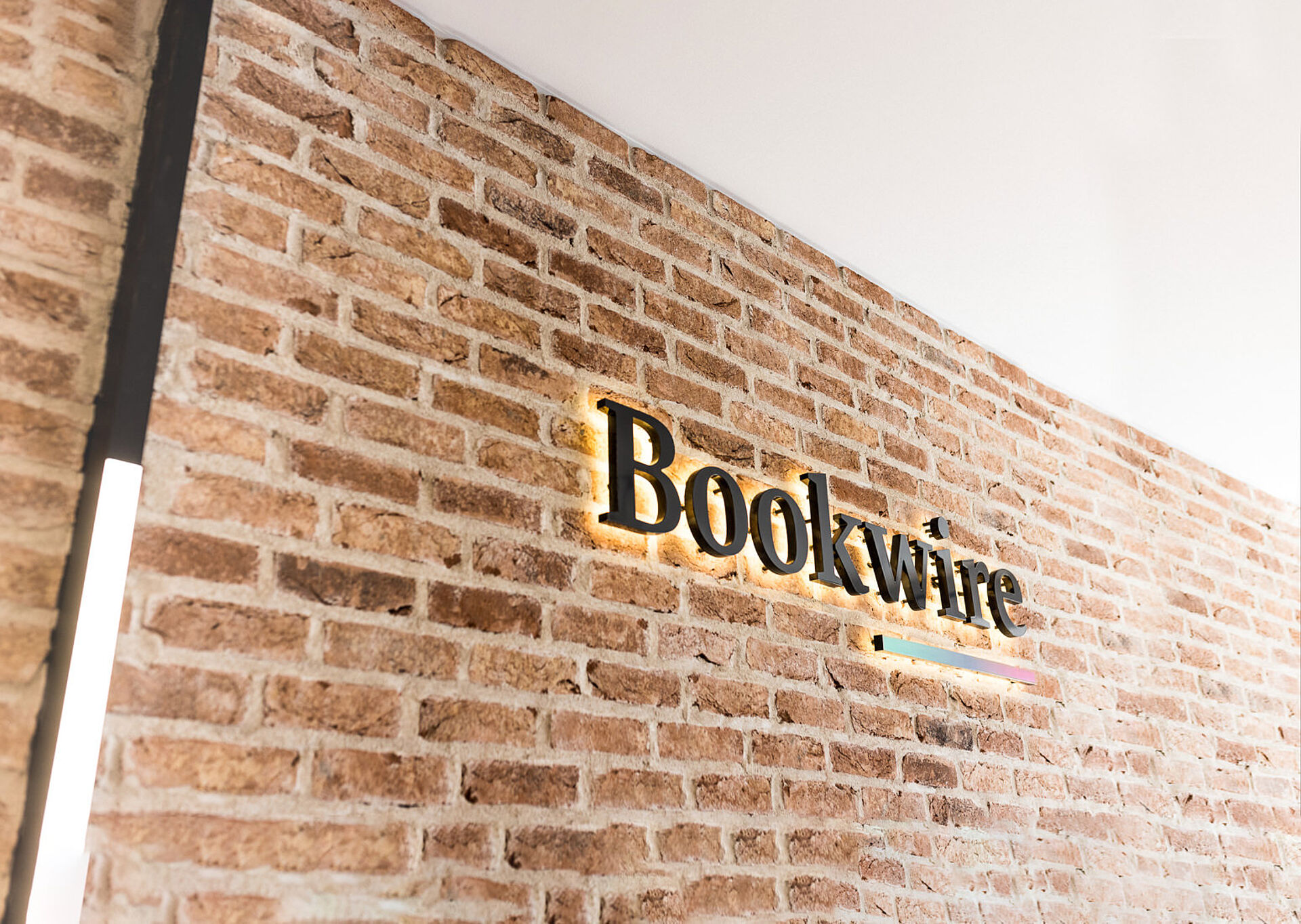 Illuminated ‘Bookwire’ lettering as a wall logo on an exposed brick wall in a modern reception area.